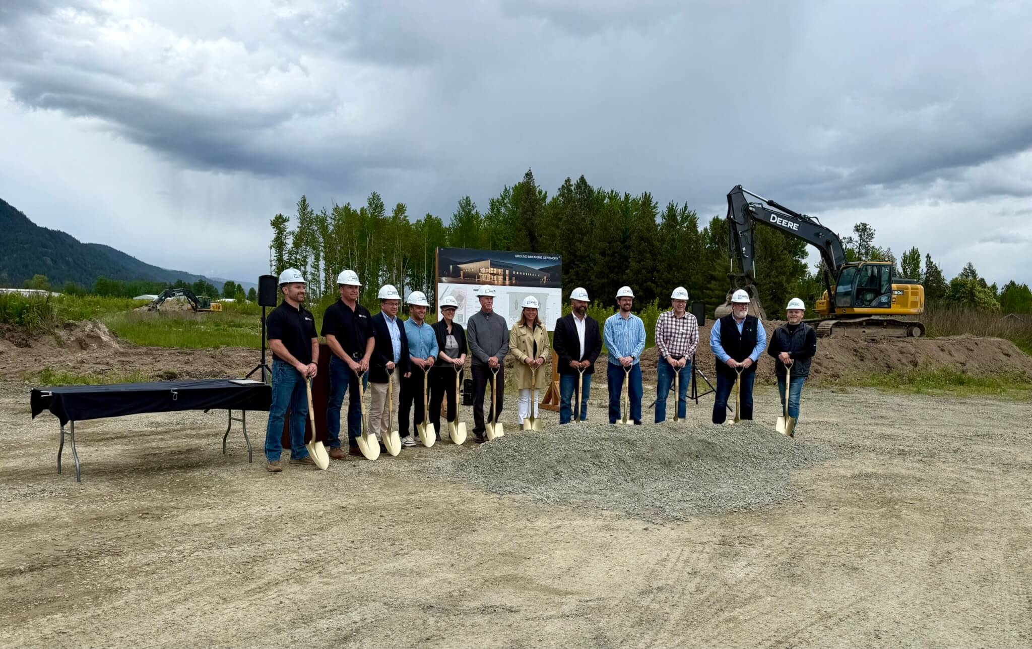 A group of people in hard hats and work clothes stand in a line holding shovels, ready to break ground on a construction site