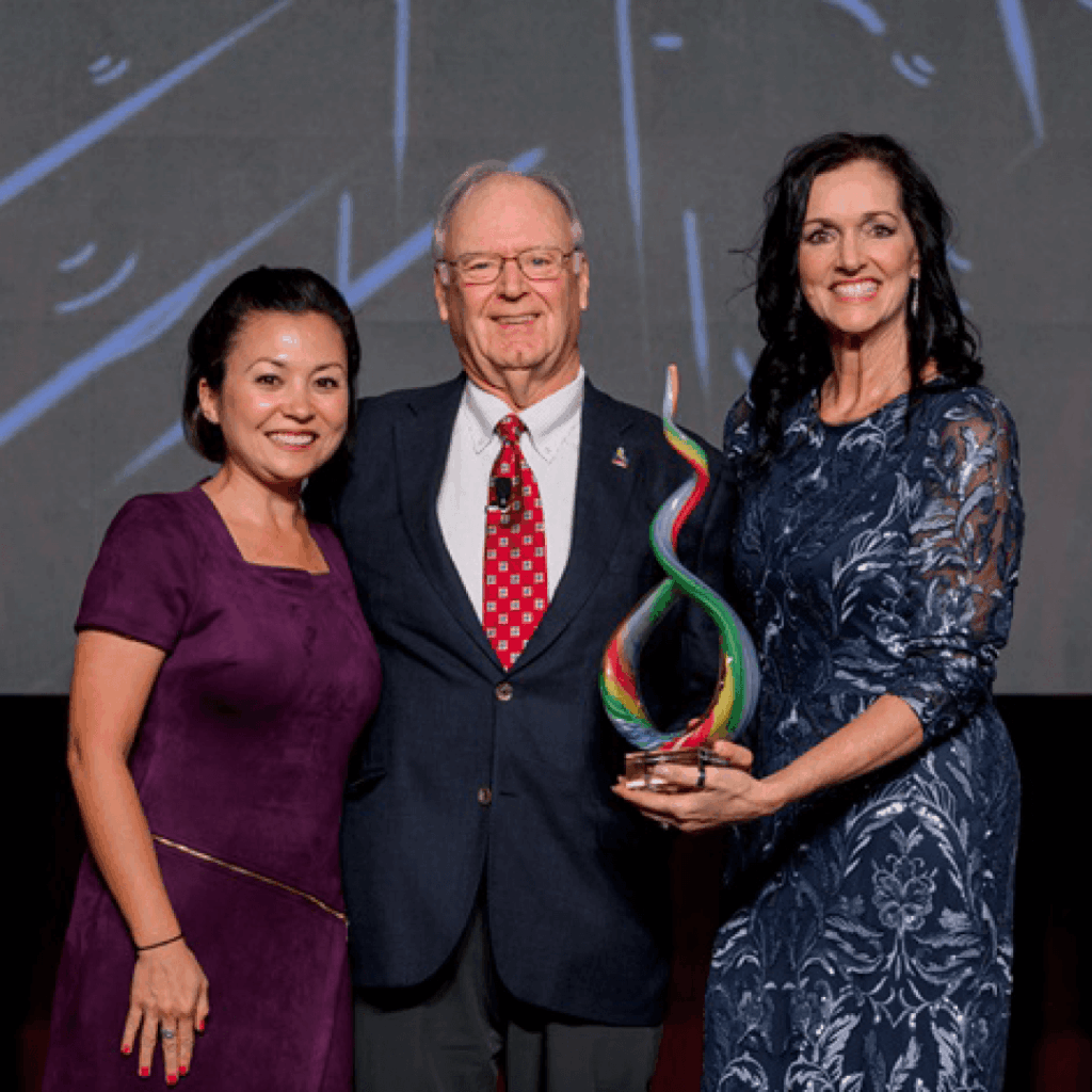 Three individuals stand together on a stage, smiling broadly as they hold a colorful, twisted glass award.