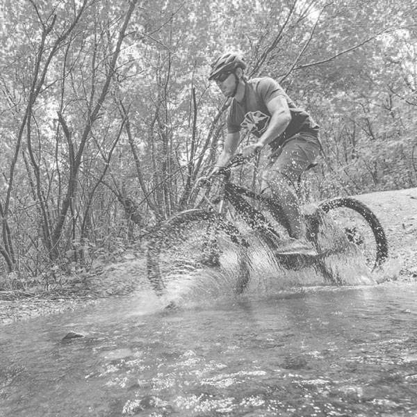 A cyclist rides through a shallow stream in a wooded area, kicking up water as they pedal