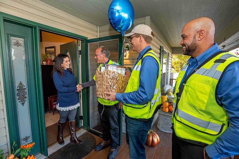 Four individuals, three wearing high-visibility vests, stand on a porch with a green door, one handing over a basket wrapped in cellophane to a smiling woman inside the doorway